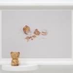 A small teddy bear figurine sits on a ledge next to a framed photo of its shattered body