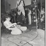 Black and white photograph of punk rock group DEVO performing at a concert In the scene several members of the group are playing guitar and bass while either jumping standing or sliding on the stage floor surrounded by sound equipment and members of the audience