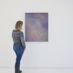 A woman stands to the left and faces a multicolored abstract painting, which hangs on a white wall.