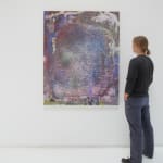 A man stands to the right and faces a multicolored abstract painting hanging on a white wall.