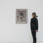 A woman admires a dark grey and white abstract painting, framed in black, hanging on a white wall.