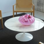 A dark-pink, cushion-like blown-glass sculpture on a white, marble coffee table.