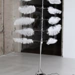 Still image of a moving mechanical sculpture made out of a long metallic pole with five different pairs of horizontal branches. At the ends of the branches white ostrich feathers catch the air currents around them