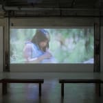 Still from video playing in gallery; a girl in the woods looks down at the animal she carries in a sling around her neck.