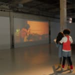 Still from video playing in gallery; a girl kneels next to a white brick wall.