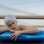 A large sculpture of a white woman in an blue innertube with a Swarovski crystal cap, a city and bridge are in the background. It's New York.