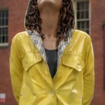 A woman in a yellow rain coat with brown hair looking up towards the sky with her eyes closed in new york city, a large, monumental sculpture