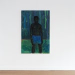 Installation view of a figurative painting by Stephen Price in a gallery setting. The canvas depicts a standing Black male figure in blue shorts, surrounded by lush green forest.