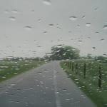 A country road leading to the horizon with grassy banks on either side of it. The right side has fencing leading along it, seen through a glass screen with water droplets scattered across it. By artist Esther Nienhuis.