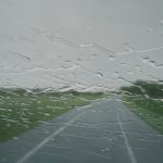 A road leading to a grey sky horizon with grassy banks on either side, seen through a glass screen with water droplets scattered across it. By artist Esther Nienhuis.