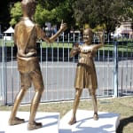 bronze sculpture of two children interacting joyfully. The boy, dressed in a casual shirt and shorts, gives a thumbs-up gesture, while the girl, wearing a dress, smiles and waves with one hand while holding a smartphone in the other, capturing the moment. The sculptures are set outdoors on a grassy area, with a metal fence and trees visible in the background.