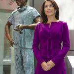 A bronze statue of Dr. Victor Chang, depicted wearing scrubs and holding a stethoscope, stands beside HRH Princess Mary of Denmark. She is wearing a vibrant purple dress with long sleeves, smiling softly, and standing with her hands clasped in front of her. The scene is set indoors with glass walls in the background.