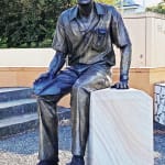 A full view of the bronze statue of Max Watters seated on a sandstone pedestal, placed outside in a courtyard surrounded by greenery and modern architectural elements. The statue captures Watters’ relaxed posture and thoughtful expression.