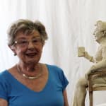 An older woman with short hair, wearing glasses, a blue top, and a silver necklace, smiling next to a clay statuette of herself holding a book and seated on a chair. The background is a plain white sheet.