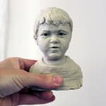 A small clay bust of a young boy with short hair, sculpted in plasticine. The figure has a soft, expressive face with slightly parted lips, capturing a sense of innocence. The bust is held in someone's hand against a plain white background.
