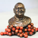 A miniature bronze bust of Lindsay Fox, mounted on a base surrounded by bright red spheres representing Jaffa lollies. The Jaffa lollies symbolize Fox's personality: hard on the outside but soft on the inside. The bust captures his distinctive features with a polished metallic finish.