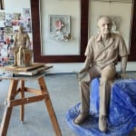 A clay sculpture of Max Watters in progress, displayed in a sculptor's studio. The life-sized figure is seated on a rock-like base with a smaller clay maquette of the sculpture placed on a wooden table beside it. Behind the scene, walls are decorated with newspaper clippings and artwork.