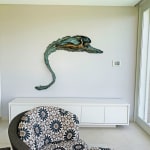 A bronze relief sculpture titled "Freestyle" installed on a wall in a bright, modern room. The swimmer's form is dynamic and surrounded by flowing aquatic textures. A patterned chair and a large glass window overlooking a pool add context to the setting.