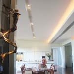 The Tinkerbell bronze sculpture mounted on a dark wall inside a modern dining area. A seated elderly man in the foreground looks toward the artwork, highlighting its prominent placement in the room.