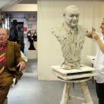 "Artist Linda Klarfeld sculpting a clay bust of Lord Alistair McAlpine in her studio in March 2012. Lord McAlpine, wearing a brown suit and colorful tie, sits nearby in a chair, engaging in conversation. The partially completed bust rests on a stand, showcasing detailed work on his facial features and attire. The studio background includes other sculptures and artistic works in progress.