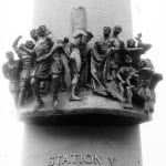 bronze relief sculpture depicting Station V of the Stations of the Cross, showing Jesus being whipped as he carries the cross. The detailed composition includes Roman soldiers in dynamic poses, some holding whips and others jeering, while Jesus endures the suffering. The scene conveys intense emotion and drama, mounted on a cylindrical structure with the inscription 'STATION V' below.
