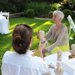 Linda Klarfeld sculpting a miniature bust of Carina in an outdoor setting. Linda is seated at a table, holding the clay bust while observing her subject, Carina, who sits across from her. Carina wears a light blouse and a yellow skirt, and the surrounding garden and lawn provide a serene atmosphere, with sculpting materials visible on the table.