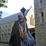 The back view of a bronze statue depicting a female graduate in flowing academic robes. She is raising her cap in celebration, with her long hair cascading down her back. The detailed folds of her gown create a dynamic and celebratory posture. The backdrop features the historic stone architecture of the Manly International School of Management, highlighting the heritage setting.