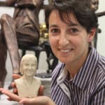 Gina holding the clay model of her sister Lisa Mari’s miniature bust in her hands. Gina is smiling warmly, with a background of bronze sculptures in an artist's studio. This miniature bust was commissioned by Gina in memory of her sister Lisa.