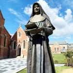 Full view of the bronze statue of St. Mary MacKillop set against a backdrop of historic church architecture. The statue, holding a book and rosary, symbolizes her contributions to education and faith.