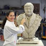 Linda Klarfeld, wearing a white coat, smiles while working on the clay model of the Dom Lopez bust in her studio. The clay bust features the likeness of Dom Lopez wearing a detailed ceremonial chain, capturing his expression with precision. The background of the studio shows art materials and tools, providing a glimpse into the sculptor’s creative process