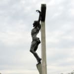 Bronze sculpture of Jesus on the cross, viewed from the side. The angled perspective highlights the realistic anatomy and the wooden texture of the cross, set against a cloudy backdrop