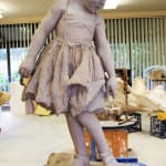 Clay sculpture in progress of a little girl who glances down, showcasing her innocence and distraction.