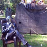 A resin sculpture titled Chairman depicting a seated male figure in a suit. The man is portrayed in a thoughtful pose, leaning back slightly in a high-backed chair with his legs crossed and hands clasped together. The figure’s head and hands have chrome finishes, contrasting with the darker patina of the suit and chair. The piece exudes authority and contemplation. The sculpture is displayed in roped off area in a park next to a large sandstone boulder which four children are sitting on with their legs dangling over the side.