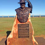 Bronze bust of Lord Alistair McAlpine mounted on a rust-colored stone pedestal, with an engraved plaque below. The sculpture features McAlpine wearing a wide-brimmed hat and a cockatoo perched on its brim. The statue is set in an open grassy area with a clear blue sky and ocean visible in the background.