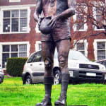 A full view of the Neil Roberts bronze statue, placed on a sleek black pedestal. The statue depicts the footballer standing tall, holding a football, with his muscular frame and poised demeanor symbolizing strength and pride. The vibrant backdrop of the lush greenery contrasts with the rich bronze finish, enhancing the statue's presence.