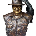 Close-up of a bronze bust of Lord Alistair McAlpine, wearing a wide-brimmed hat and a textured button-up shirt. A detailed black cockatoo perches on the brim of the hat, adding a unique element to the sculpture. The bust captures a warm and smiling expression