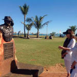 Bronze bust of Lord Alistair McAlpine on a rust-colored stone pedestal with an engraved plaque. Two women stand nearby, one gesturing toward the sculpture. The scene is set in a park with palm trees, a green lawn, and the ocean visible in the background under a bright blue sky.