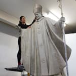 Linda Klarfeld sculpting a life-size clay model of a statue depicting Pope John Paul II in progress. The figure is shown wearing papal robes and a mitre, holding a staff topped with a crucifix in one hand and extending the other in a gesture of blessing. The sculpture is mounted on a platform covered with a blue tarp, inside a workshop with a plain white wall and fluorescent lighting