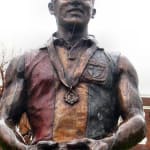 A close-up view of the Neil Roberts bronze statue, showcasing his confident facial expression. The polished surface highlights intricate details, including the medal around his neck and the texture of his uniform.