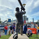 A bronze statue of Australian cricket legend Allan Border captured from behind, showing him mid-action, swinging his cricket bat high in the air. The sculpture is installed on a sandstone base at The Gabba in Brisbane, with a blue sky and event attendees in the background.