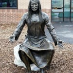 Front view of a bronze statue of Jesus seated on a rock, arms open in a welcoming gesture, with a brick building in the background.