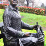 Bronze statue of Bill Woodfull, a legendary cricketer, seated on a bench holding a cricket bat, with protective gear on his legs. The detailed expression and attire honor his contributions to the sport.