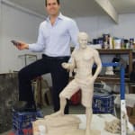 A man, Scott, is standing in a studio next to a clay sculpture modeled in his likeness. The sculpture portrays Scott in a casual pose, wearing shorts and standing barefoot on a rocky base, holding a small object in his right hand. The man smiles proudly beside his sculpted figure.