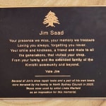 A memorial plaque dedicated to Jim Saad, titled "Jim Saad," featuring a poetic tribute to his memory and kindness. The inscription reads, "Your presence we miss, your memory we treasure, Loving you always, forgetting you never. Your smile and kindness, a friend and mate to all the generations, that visited your shop. From your family and the extended family of the Kirribilli community and beyond. Vale Jim." Below the tribute, an additional note explains that several of Jim's shoe repair tools and a pair of his boots were donated by his family to North Sydney Council in 2023. It mentions that these items were used by artist Linda Klarfeld as inspiration for the memorial sculpture. The plaque includes an emblem of a cedar tree at the top.