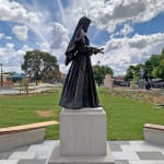 ide profile of the bronze statue of St. Mary MacKillop, displayed on a pedestal in a landscaped outdoor setting. The composition highlights her contemplative pose and enduring legacy