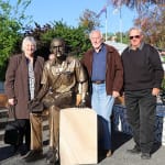 A bronze statue of Max Watters seated on a sandstone pedestal, with his left hand resting on his lap and his right hand on the pedestal. He is dressed casually, wearing a shirt with a pen in the pocket, trousers, and shoes. Surrounding the statue are three individuals: two men and a woman, all standing and smiling in a sunny outdoor setting.