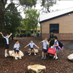 School children interact with a bronze statue of Jesus seated on a rock, surrounded by natural landscaping and large stones. The children are smiling and playing, with some sitting on the rocks and others standing, creating a lively and joyful scene. The statue is located near a brick school building amidst tall trees.