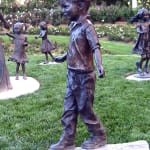 Bronze sculpture of a young boy balancing on a log, part of a whimsical children's garden scene in a lush park setting, with other bronze statues of children playing in the background.