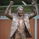 A close-up view of a bronze statue of Nick Columb standing on a green lawn, with both arms raised in a victorious gesture. He is depicted in a suit and tie, with an exuberant expression, holding a book in one hand. The statue is set outdoors, with a wooden stable-like structure in the background, symbolizing his achievements in the horse racing industry.
