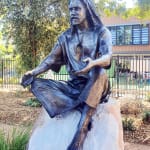 A side profile of the bronze statue of Jesus seated on a rock, with one hand extended and an inviting posture, surrounded by trees in a peaceful garden setting.